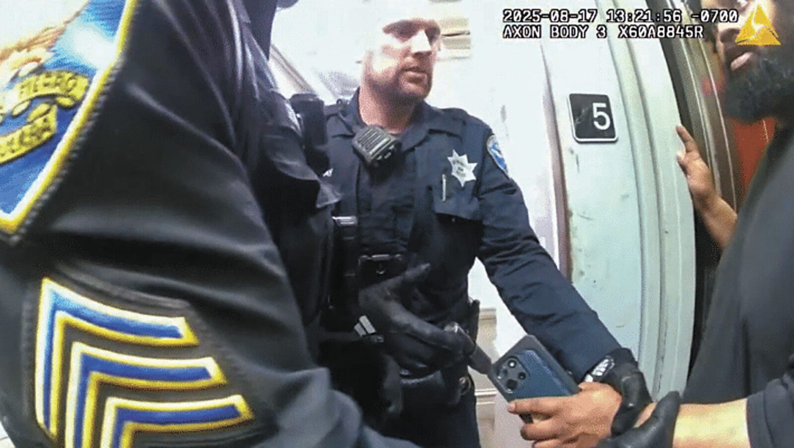 A police officer in uniform stands near a door marked with the number 5, holding a phone, while another officer in uniform is partially visible nearby.